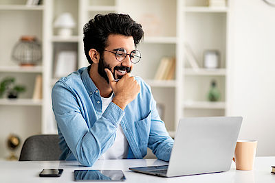 Telecommuting Concept. Smiling Indian Man Working Online With Laptop At Home Office